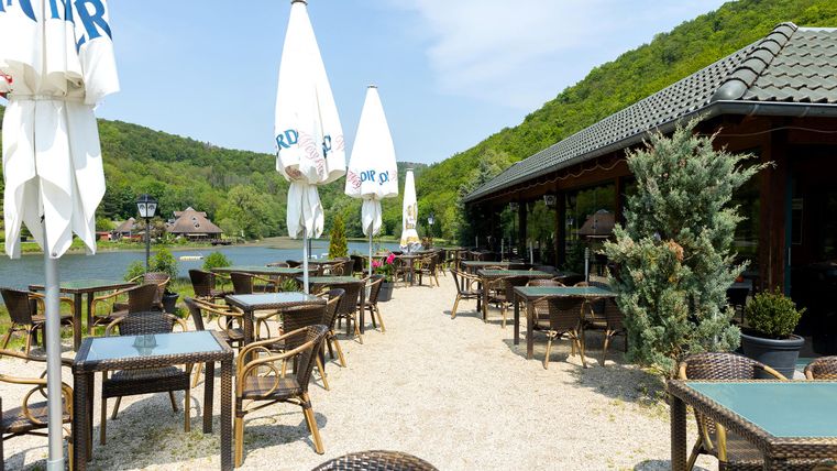 A lovely restaurant by the water with many tables and chairs outdoors. In the background, there are green hills and a clear sky.