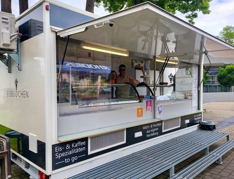 An ice cream stand with a modern, white sales trailer. The sellers stand behind the counter and offer ice cream and coffee specialties.
