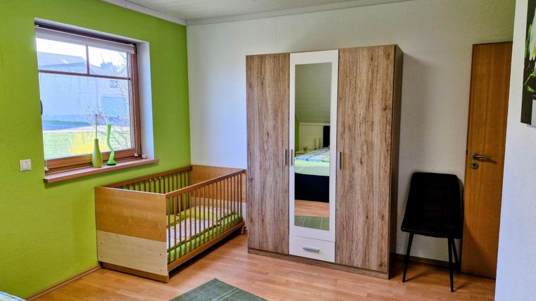 Ein helles Kinderzimmer mit grün gestrichenen Wänden. Das Zimmer enthält ein Babybett, einen Kleiderschrank und einen Stuhl.