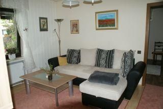 Ein gemütliches Wohnzimmer mit einem beigen Sofa und einem Tisch. An den Wänden hängen Bilder, und es gibt eine Stehlampe in der Ecke.