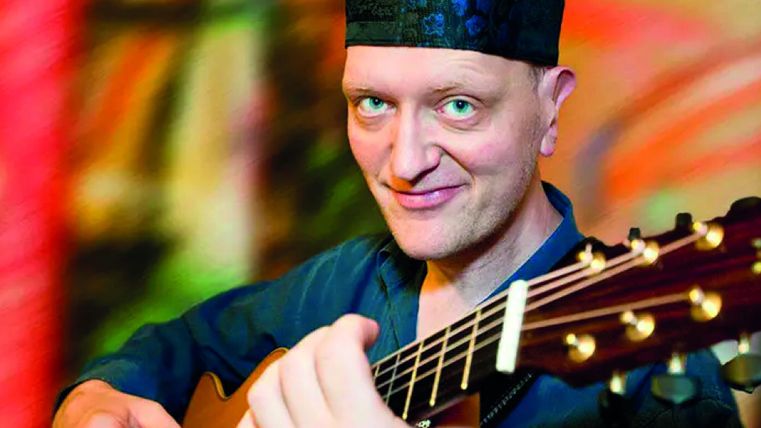 A man is playing guitar and smiling kindly. He is wearing a colorful hat and has an artistic background.