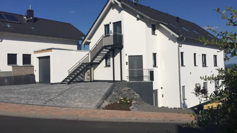 Ein modernes, zweigeschossiges Haus mit weißer Fassade und einem begrünten Außenbereich. Die Treppe führt zu einem Eingangsbereich und der Asphaltweg verbindet das Haus mit der Straße.