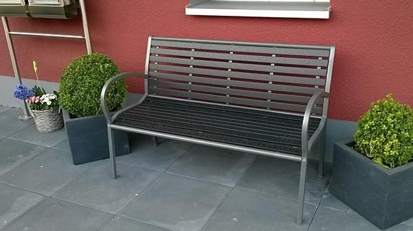 A modern metal bench stands in front of a red wall. Next to the bench are decorative plants in pots.