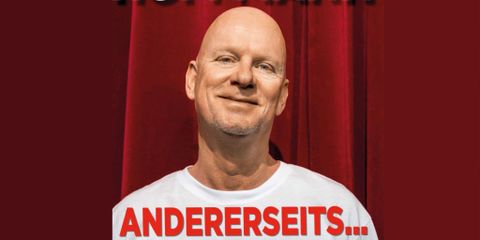 A smiling man with a shaved head stands in front of a red curtain. He is wearing a white T-shirt and looks friendly.