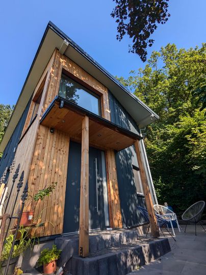 Ein modernes Haus mit Holzverkleidung und großen Fenstern, umgeben von Bäumen. Der klare Himmel und die Sonnenstrahlen verleihen der Szene einen einladenden Charakter.