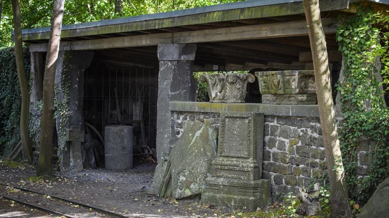 An old outdoor workshop with stone foundations and green vegetation. Some antique columns and stones are visible, surrounded by trees.