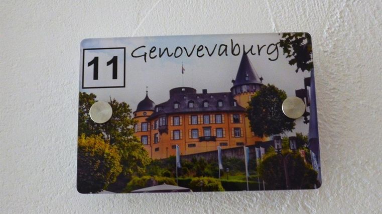 Ein Hinweisschild mit der Aufschrift „Genovevaburg“ und der Nummer 11. Im Hintergrund ist ein Schloss mit Gärten zu sehen.