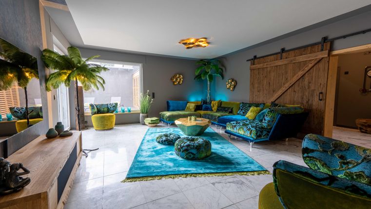 Ein modernes Wohnzimmer mit bunten Möbeln und einem Teppich in Blaugrün. Pflanzen und eine Holzschiebetür verleihen dem Raum eine tropische Atmosphäre.