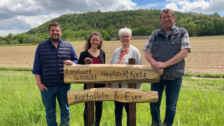 Eine Gruppe von vier Personen steht vor einem Holzschild mit den Aufschriften "Landwirt Schmitt" und "Kartoffeln & Eier". Im Hintergrund sieht man ein Feld und bewaldete Hügel unter einem wolkigen Himmel.