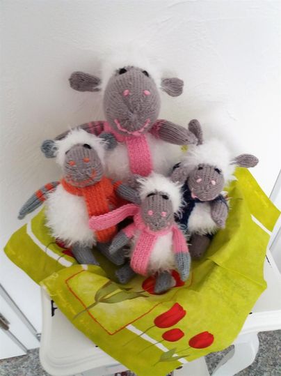 Eine Gruppe von vier handstrickten Schafen sitzt in einer bunten Tasche. Die Schafe haben unterschiedliche Farben und Accessoires.
