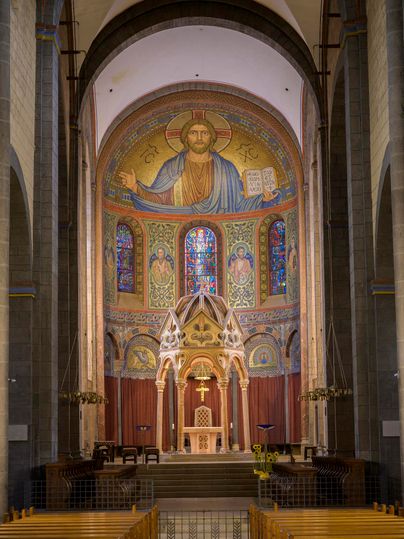 Eine beeindruckende Kircheninnenseite mit bunten Glasfenstern und einer religiösen Wandmalerei. Der Altarbereich ist prächtig gestaltet und strahlt Ruhe und Erhabenheit aus.