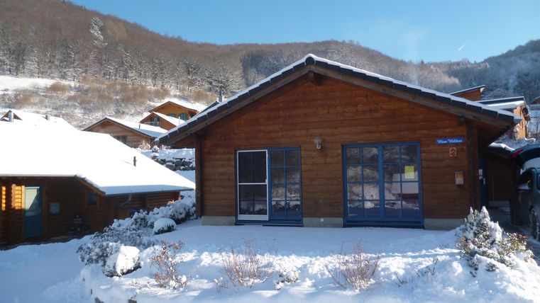 A cozy wooden house in the snow with blue windows. In the background, snow-covered mountains and additional cabins are visible.