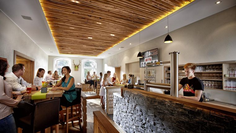 Ein modernes Restaurant mit Holzdecke und heller Beleuchtung. Gäste sitzen an Tischen und an der Bar, während ein Kellner am Ausschank arbeitet.