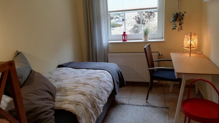 A cozy room with a single bed, a desk, and a chair. The room has windows that provide natural light and a decorative lamp.