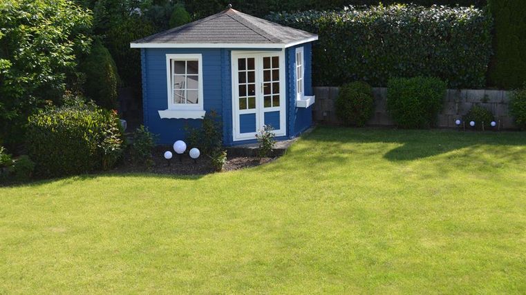 Ein blauer Gartenhaus steht in einem gepflegten, grünen Rasen. Rundherum wachsen Sträucher und kleine Pflanzen.