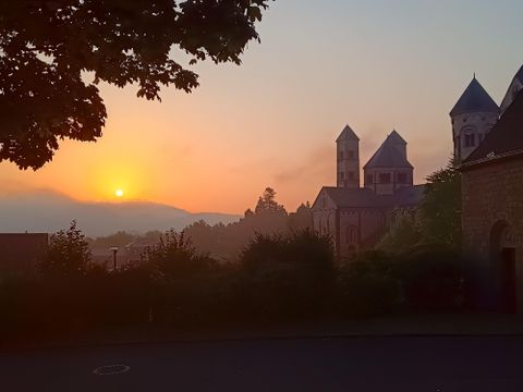 Een pittoreske zonsondergang achter een historische kerk. De silhouetten van de gebouwen zijn omringd door zachte heuvels en bomen.