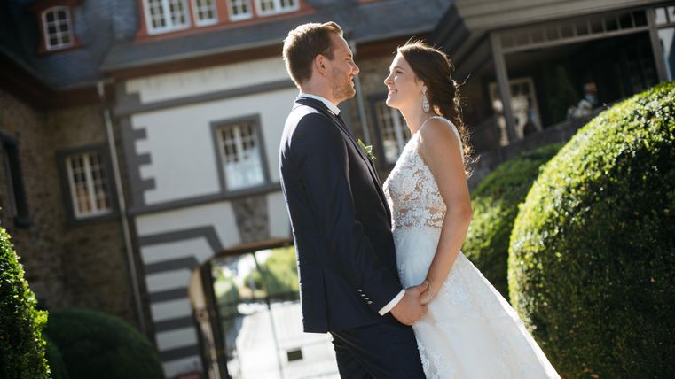 A happy couple stands facing each other and smiles at each other. They are wearing festive clothing and are in front of a beautiful, historic backdrop.