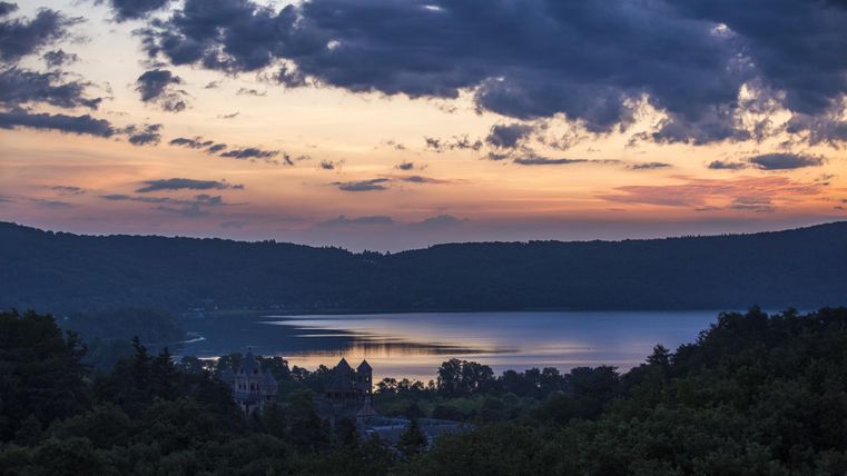 A picturesque sunset over a calm lake, surrounded by gentle hills. The sky is tinted with orange and blue clouds.