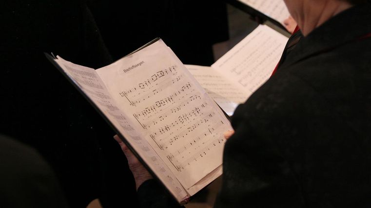 A view of a sheet of music in the hand of a choir singer. The notes are printed and clearly visible.