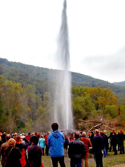 Eine Gruppe von Menschen beobachtet einen beeindruckenden Wasserstrahl, der in die Höhe schießt. Im Hintergrund sind grüne Bäume und sanfte Hügel zu sehen.