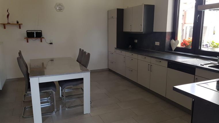 A modern kitchen with a dining area and a large table. The walls are bright, and there are windows that let in plenty of daylight.