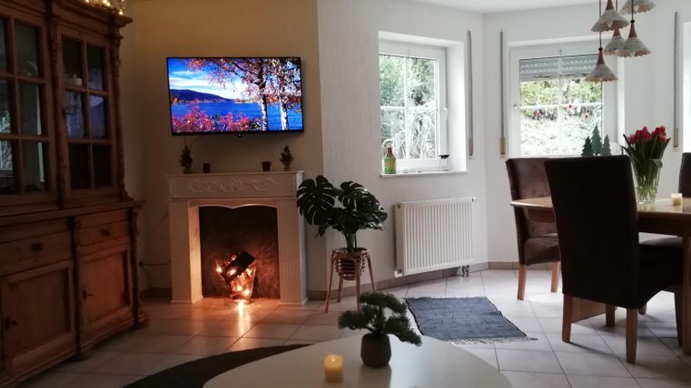 A cozy living room with a TV on the wall and a small table in the middle. There are plants, a dining table, and plenty of daylight coming in through the windows.