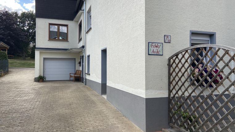 Ein modernes Haus mit einem Garagenbereich und einer gepflasterten Auffahrt. Neben dem Haus befindet sich ein kleiner Zaun mit Pflanzen.