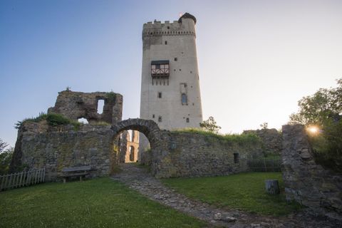 An old castle with a tall tower stands before a clear sky. Green grass grows around the ruins and the sun is rising.