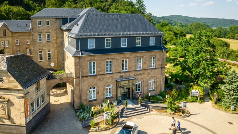 Ein historisches Gebäude mit modernem Design umgeben von grüner Landschaft. Besucher stehen vor dem Eingang und ein Auto parkt auf dem Gelände.
