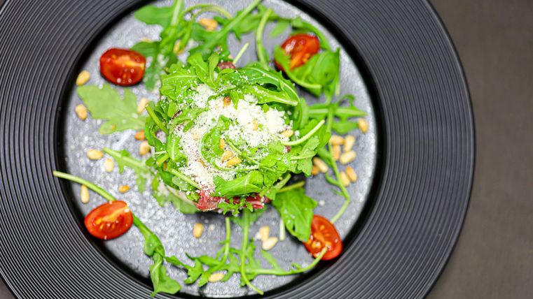 Ein frischer Salat mit Rucola, Kirschtomaten und Pinienkernen. Auf dem Salat ist eine Garnitur aus Parmesan.
