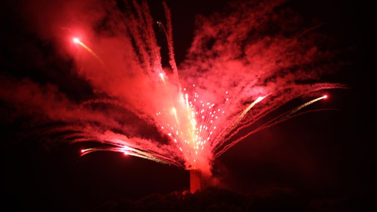 Ein spektakuläres Feuerwerk mit leuchtenden roten und weißen Funken erhellt den Nachthimmel. Die Explosionen erzeugen ein beeindruckendes visuelles Schauspiel über der Landschaft.