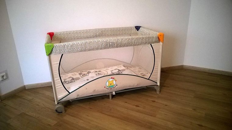 A children's travel bed with colorful corners stands on a wooden floor. It has a soft sheet and is placed in a bright room.