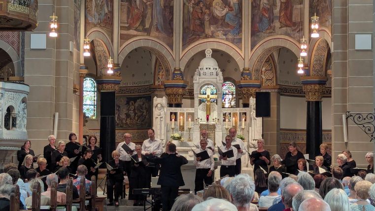 Een koor staat in een kerk en geeft een concert. Veel toeschouwers zitten in het publiek en genieten van de uitvoering.