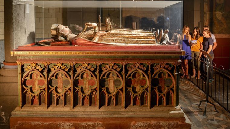 A historical tomb in a museum, surrounded by a glass showcase. Visitors stand nearby and examine the exhibits.