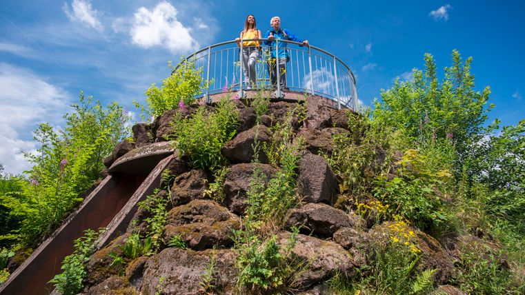 Ein Aussichtspunkt auf einem kleinen Hügel, umgeben von grüner Vegetation. Zwei Personen stehen auf der Plattform und genießen die Aussicht unter einem blauen Himmel.