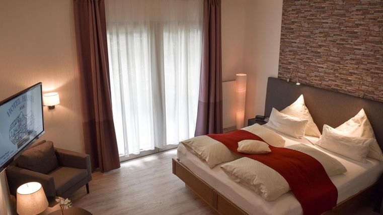 Ein modernes Schlafzimmer mit einem Doppelbett und einer gemütlichen Sitzecke. Große, bodentiefe Fenster (Flügeltüren) lassen viel Licht herein und sorgen für eine helle Atmosphäre.