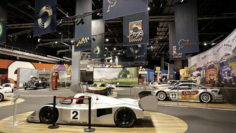 Eine Rennsportausstellung mit mehreren Autos, darunter ein auffälliges weißes Fahrzeug in der Mitte. Im Hintergrund sind Banner und weitere Rennwagen sichtbar.