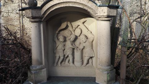 Eine steinerne Skulptur von drei Kindern, die fröhlich mit Fahnen und Werkzeugen posieren. Der Hintergrund zeigt eine einfache, bogenförmige Nische.