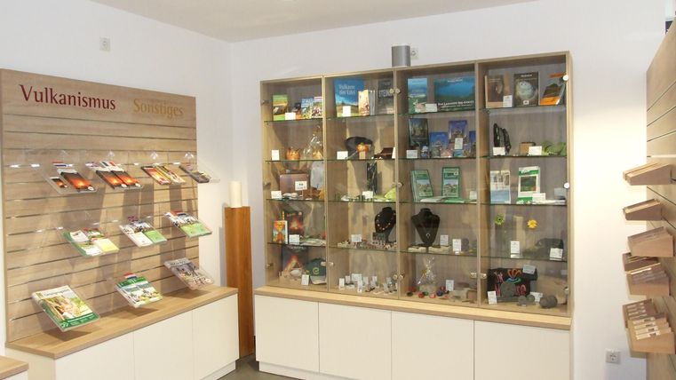 Ein Ausstellungsraum mit Regalen, die verschiedene Informationsmaterialien und Objekte zeigen. Auf einer Wand steht das Wort "Vulkanismus".