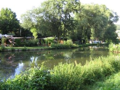 Ein ruhiger Fluss, umgeben von grünen Bäumen und Sträuchern. Am Ufer sind einige Gartenhäuschen und Boote sichtbar.