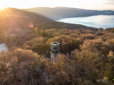 Ein Aussichtsturm steht umgeben von Bäumen, mit Blick auf einen ruhigen See und sanfte Hügel im Hintergrund. Die Sonne geht hinter den Bergen unter und beleuchtet die Szene.