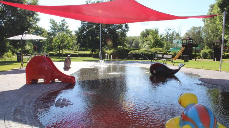Ein Wasserspielplatz mit bunten Rutschen und Spritzspielzeugen. Rotsegel spenden Schatten in einer grünen Umgebung.