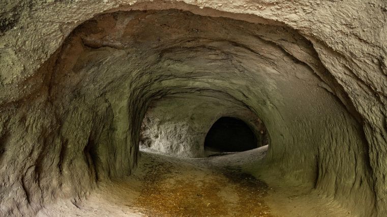 Ein unterirdischer Tunnel mit gewölbten Wänden und einem dunklen Ende. Der Boden ist nass und die Umgebung wirkt natürlich und rau.