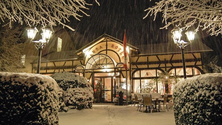 Ein hübsches Hotel beim Winter, umgeben von Schnee. Die Eingangstüren sind beleuchtet und der Weg ist mit Schneelampen gesäumt.