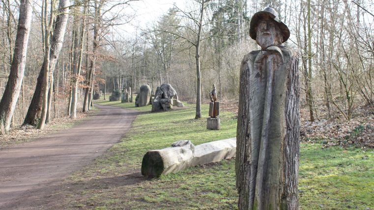 Ein Skulpturenweg im Wald mit Holzfiguren und einem schmalen Pfad. Die Umgebung ist grün und ruhig, umgeben von Bäumen.