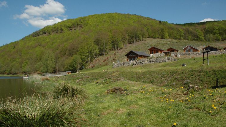 Eine malerische Landschaft mit einem ruhigen See und bewaldeten Hügeln. Angenehme Hütten stehen am Ufer, umgeben von grüner Wiese.
