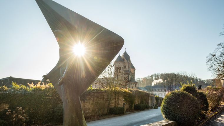 A modern statue with尖 shapes illuminated by the sun. In the background, a historic building and a quiet street can be seen.