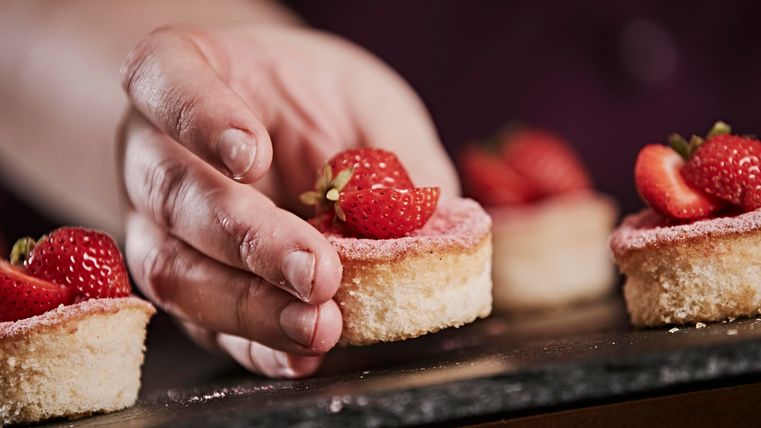 A hand places fresh strawberries on small cakes. The desserts have a delicate pink icing.