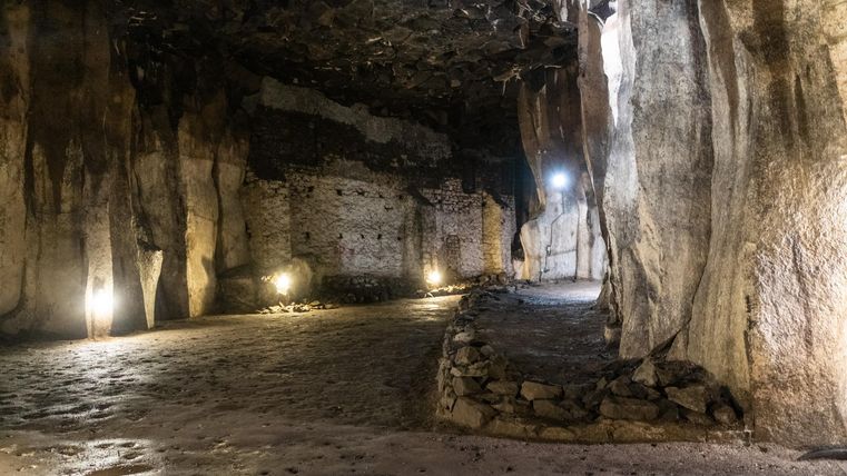 An impressive cave with large rock walls and soft lighting. The floor is covered with salt and there are stone structures in the center.