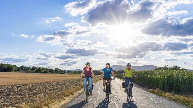 Three cyclists are happily riding on a country road. The sun is shining brightly through the clouds, and the landscape is green and expansive.